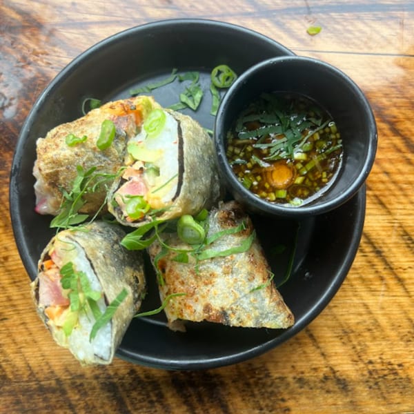 Crispy fried spring rolls with fresh herbs and a side of dipping sauce on a black plate.