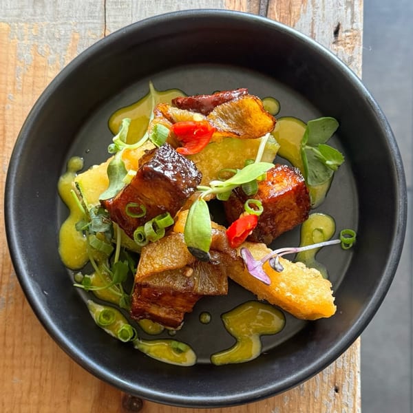 Crispy pork belly with green sauce, red peppers, and microgreens in a black bowl.