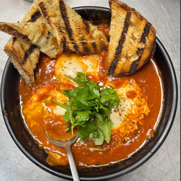 Shakshuka with two fried eggs, fresh greens, and grilled bread in a black bowl.