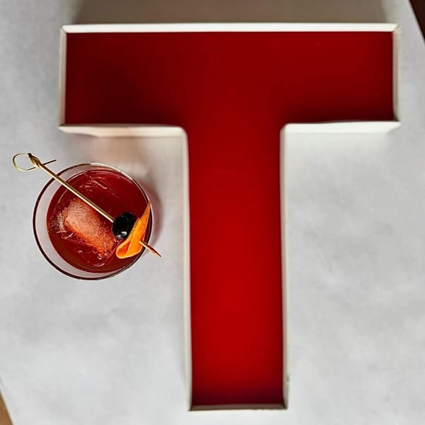 A red cocktail with an orange peel and cherry next to a large red letter T.